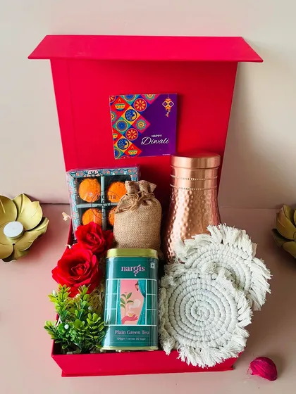 This premium red flap-open box contains a copper bottle, macrame coasters, green tea, almonds, and unique laddoo-shaped candles.
