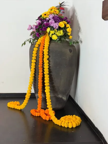 A decorative corner piece featuring a dark earthen pot overflowing with colorful flowers and bright marigold garlands. This simple arrangement adds a touch of traditional charm to any festive space.