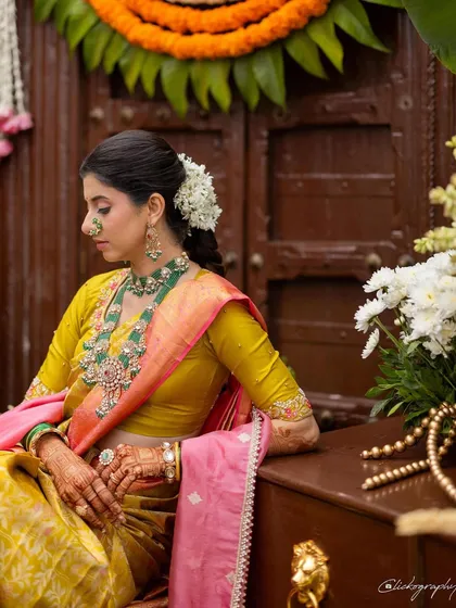 A quiet, contemplative portrait of the bride. The styling focuses on traditional details like the gajra in her hair, the classic nath, and the intricate design of her emerald necklace.