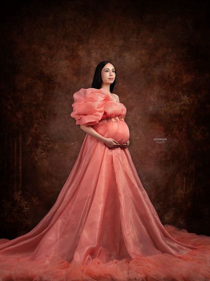 A full-length portrait showcasing a stunning peach gown with a dramatic, single ruffled sleeve. The dark, painterly background makes the color of the dress pop, creating a beautiful fine art piece.