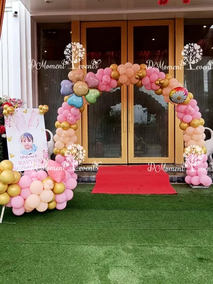 A charming balloon arch entrance for a first birthday party. The pink, gold, and multicolored balloons create a festive welcome, complemented by a personalized welcome stand featuring the birthday girl's photo.