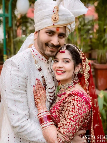 A classic wedding portrait of the smiling couple. Both are looking at the camera, radiating happiness, showcasing their coordinated red and white traditional outfits in a beautiful garden setting.