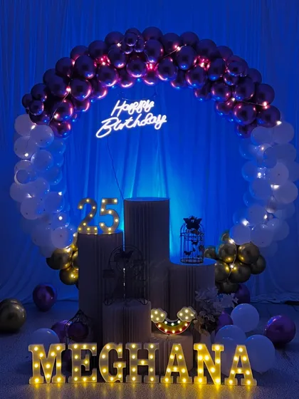 A 25th birthday celebration with a moody and stylish theme. The circular balloon arch uses purple, white, and gold balloons, with a neon sign and marquee letters for 'MEGHANA' creating a beautiful glow.