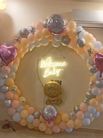 A classic "Welcome Baby" decoration. This full balloon ring in pastel peach, pink, and silver, with a neon sign and baby-themed foil balloons, is perfect for a baby shower or welcome party.