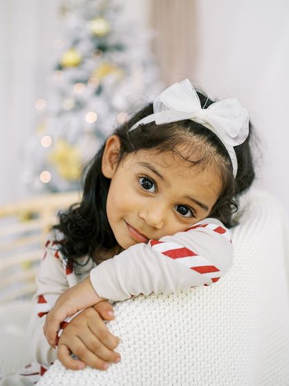 A close-up of a little girl with a sweet expression, with the twinkling lights of the Christmas tree in the background.