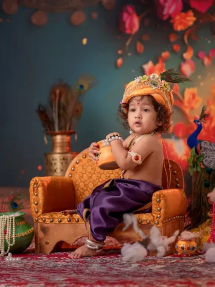 A baby dressed as Krishna sits on a miniature throne, holding a small pot of butter. The rich details, from the pearl necklaces to the peacock feathers, make this a standout portrait.