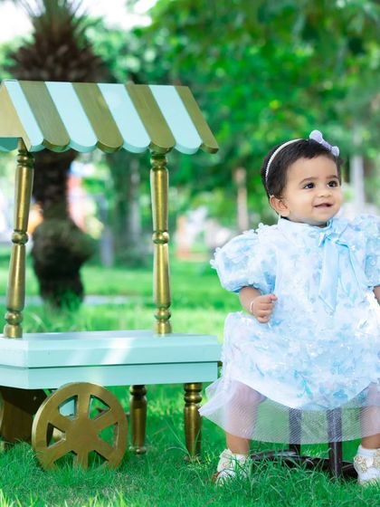 A sweet baby girl posing with a miniature ice cream cart in a beautiful green park.