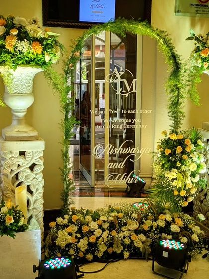 A personalized welcome mirror for an engagement ceremony, etched with the couple's monogram and a sweet message. The arch is framed with fresh greenery and white and peach florals, creating a charming entrance.