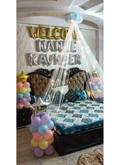 A sweet and personal welcome home setup over the bed. It features a canopy with fairy lights, "Welcome Nanke Kavneer" foil balloons, and colorful balloon pillars.