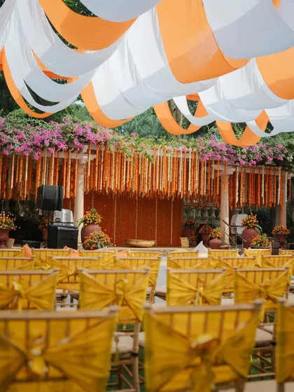 A wide view of the Haldi ceremony setup, featuring orange and white drapes, marigold garlands, and bright yellow seating for guests.