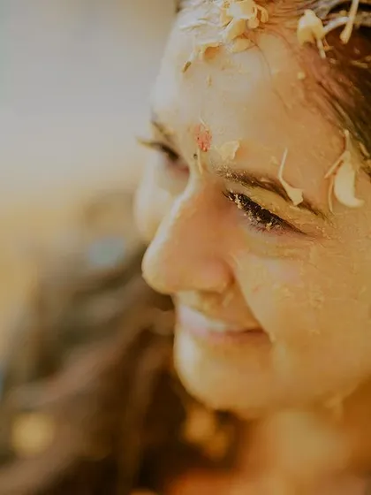 A close-up shot capturing the bride's serene smile during her haldi. Covered in turmeric paste and flower petals, she radiates a natural glow and happiness.
