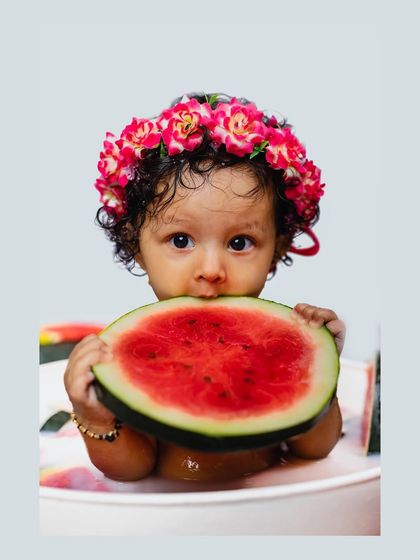 A baby girl holds a large slice of watermelon up to her mouth while sitting in a milk bath. This adorable photo perfectly encapsulates the fun of a themed baby shoot.
