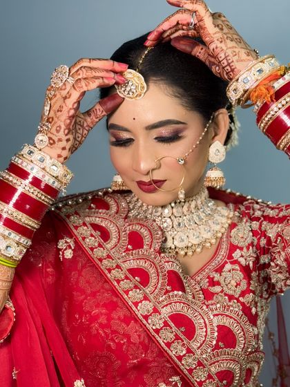 A beautiful bride adjusting her maang tikka. She is wearing a red lehenga with a contrasting dupatta, and her makeup features a soft pink and red eye look.