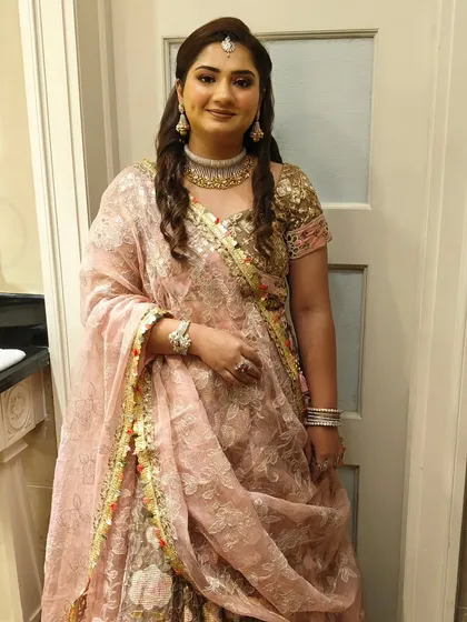 A full-length view of a stunning party look for a wedding event. The hair is styled with voluminous waves and a side part, perfectly matching her traditional pink lehenga.