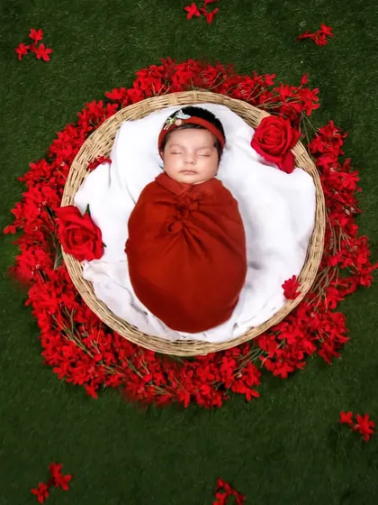 A vibrant overhead shot of a baby in a red wrap, nestled in a basket surrounded by a ring of bright red flowers on a green lawn.