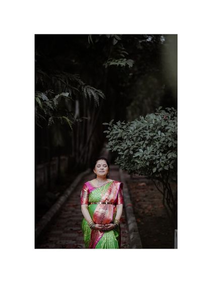 A serene portrait of an expecting mother during her baby shower. Dressed in a beautiful saree, she stands on a path surrounded by greenery, with her eyes closed in a moment of peace.