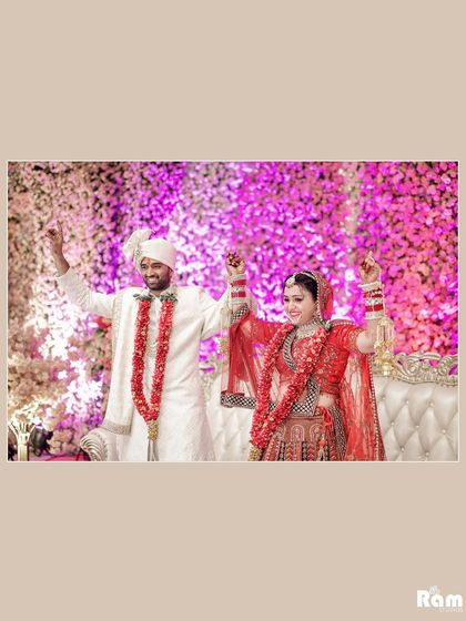 A joyful shot of the couple on their wedding stage, hands raised in celebration against a backdrop of purple flowers.