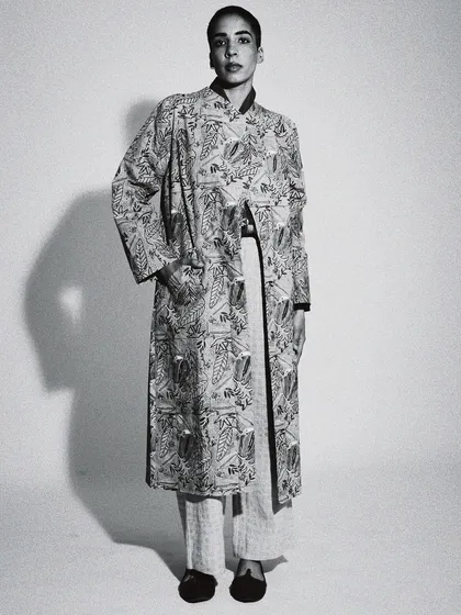 A full-length black and white portrait with strong, graphic lighting. This image has a minimalist, high-fashion feel, focusing on the silhouette of the long jacket.