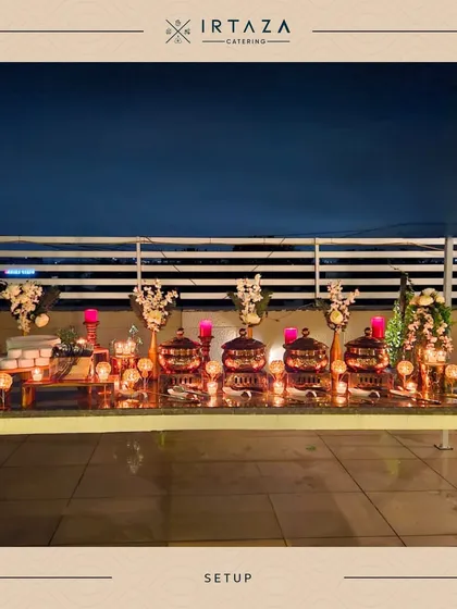 A wide shot of a rooftop dinner setup against the evening sky. The long line of glowing chafing dishes and candles creates a stunning visual and promises a memorable dining experience.