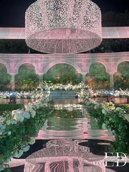 The reflection of the grand chandelier on the glossy black aisle creates a dramatic and mesmerizing effect. The path is flanked by abundant white floral arrangements, guiding the way to the main stage.