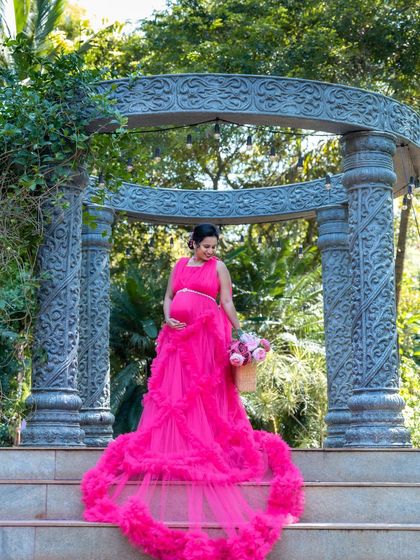 A stunning shot of a mom-to-be in a bright pink ruffled gown, standing on the steps of a stone archway. She is holding a basket of flowers, looking down at her bump.