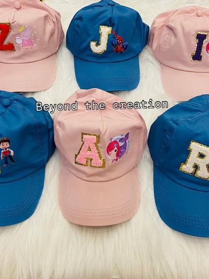 An overhead view of several personalized caps ready for a party. Each cap, in either blue or pink, features a unique combination of an initial and a character patch, from Peppa Pig to superheroes.