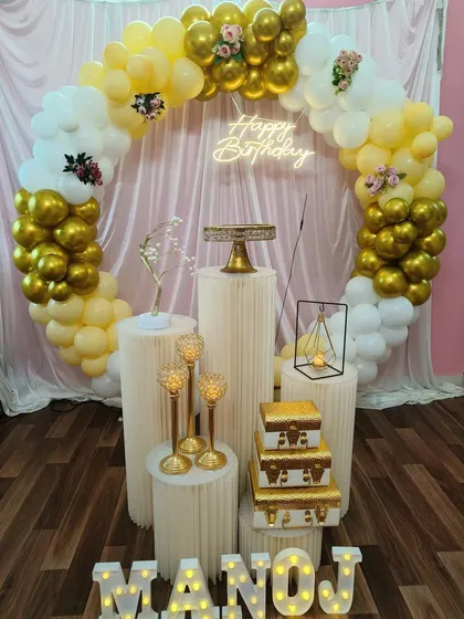 A warm and sunny yellow, white, and gold balloon ring for Manoj's birthday. The decoration is complemented by gold candle holders and a light-up name sign.