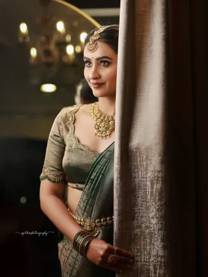 A candid moment captured with artistic flair. Peeking from behind a curtain, the bride's smile is genuine, and the soft lighting from the chandelier in the background adds a touch of glamour.