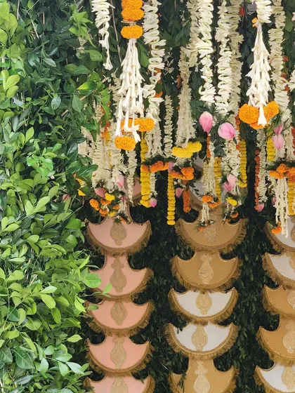 A detail shot of a South Indian mandap backdrop. A wall of lush green leaves is adorned with hanging floral strings and traditional fabric fans, creating a rich tapestry of textures and colors.