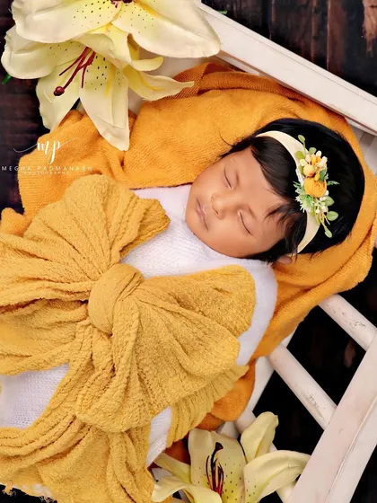 A close-up of the baby's face, framed by the soft yellow wrap and lily petals.