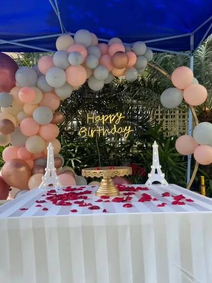 A romantic birthday cake table setup under a canopy, featuring a pastel balloon arch and a neon "Happy Birthday" sign.