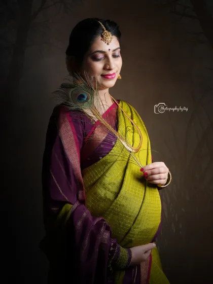 An artistic studio portrait of a mother-to-be in a traditional saree, holding a peacock feather. The lighting and pose evoke a sense of timeless beauty.