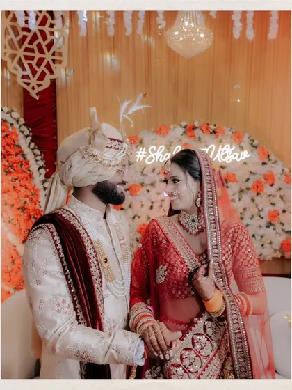A beautiful portrait of Shalini and Utsav on their wedding day, standing before a floral wall with their wedding hashtag lit up.