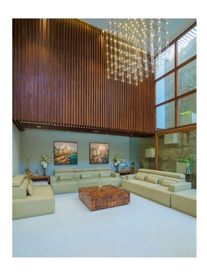 The double-height living area in "Athulyam" is a voluminous space enhanced by natural light from large windows. Vertical wooden slats and a dramatic chandelier add scale and texture.