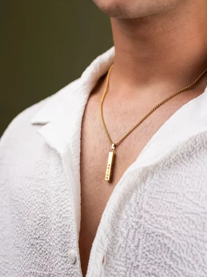 This lifestyle shot shows how a piece of jewelry complements personal style. The focus is on the gold pendant against a simple white shirt, demonstrating the product in a natural, wearable context for a men's accessory brand.