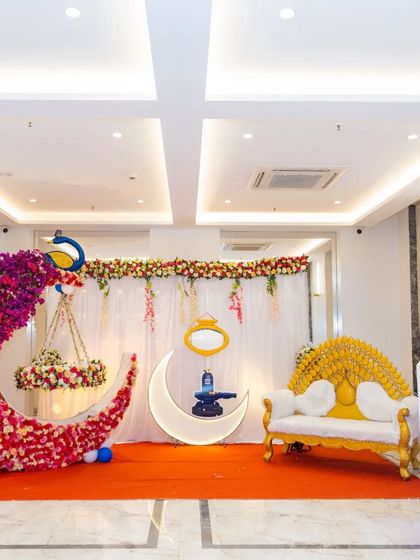 A grand naming ceremony decoration with a large floral moon cradle topped with a peacock. The golden throne-style sofa and 'BABY BOY' letter blocks complete this royal setup.