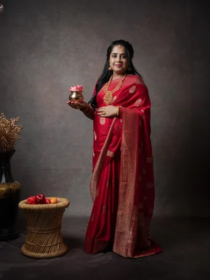 A full-length studio portrait for a traditional baby shower or Sreemantha ceremony. The setup includes classic props to complement the rich red saree and temple jewelry.