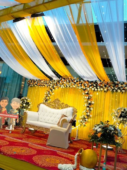The Haldi stage viewed from the front, with its bright yellow backdrop, floral arch, and elegant white sofa.