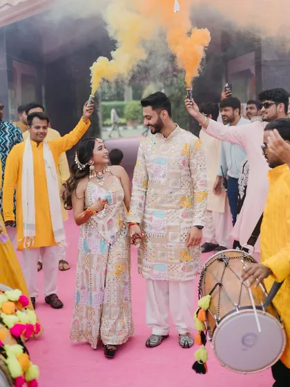 The couple makes a grand entrance to their Haldi ceremony, walking hand-in-hand through a cloud of yellow smoke bombs while a dhol player leads the way.