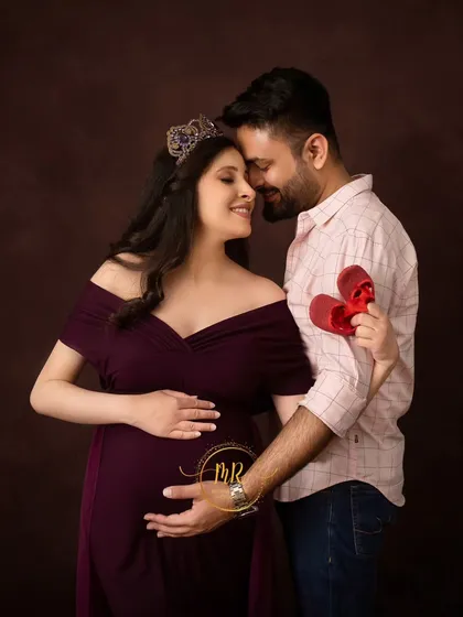 A cute and symbolic couple's portrait. The partner holds up a tiny pair of red baby shoes while embracing his wife, who is wearing a beautiful crown and purple gown.