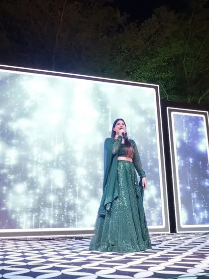 Dressed in a beautiful green lehenga, I'm addressing the audience at an outdoor Sangeet, with a stunning LED backdrop lighting up the night.