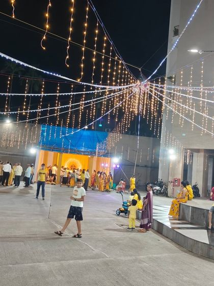 A wide shot of a Navratri celebration in a society compound. The area is beautifully lit with strings of fairy lights, creating a magical atmosphere for the garba night.