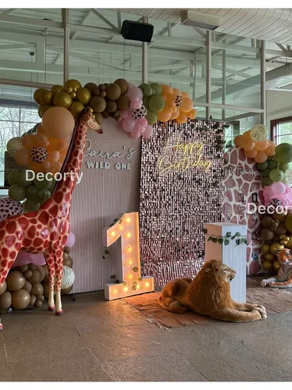 A chic "Wild One" jungle safari theme. The backdrop includes a rose gold sequin wall, a neon "Happy Birthday" sign, and life-sized giraffe and lion props for a glamorous touch.