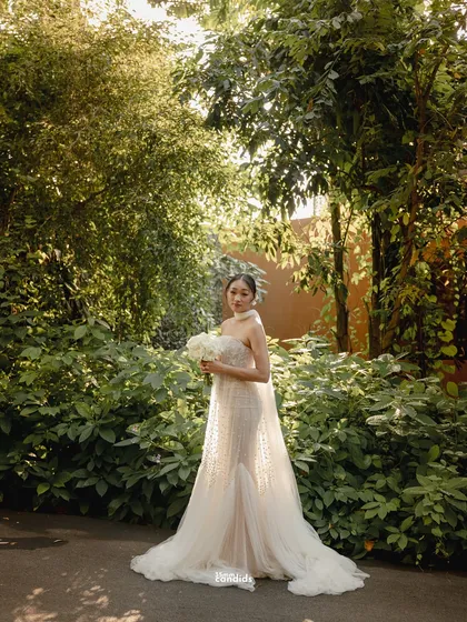 A stunning full-length bridal portrait in a lush garden setting. The bride holds her bouquet, her long white dress flowing behind her, creating a picture of pure elegance.