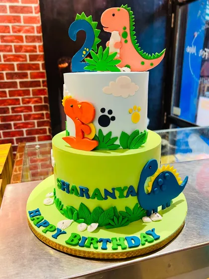A colorful two-tier dinosaur cake for Sharanya's 2nd birthday, with friendly fondant dinosaurs and edible paw prints.