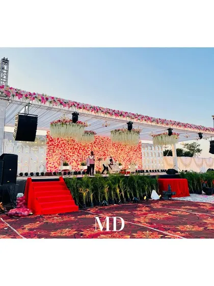 A daytime view of a large outdoor wedding stage setup. This image shows the scale of the structure, with its tall support pillars, extensive floral arrangements, and professional sound system integration.