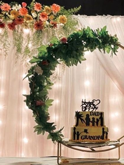 I designed this beautiful cake display for a special family celebration. A golden crescent-shaped frame adorned with greenery and flowers creates an elegant stand for the cake, set against a backdrop of fairy lights.