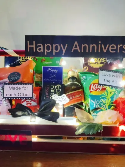 An anniversary hamper illuminated with fairy lights to set a romantic mood. It includes a bottle of Red Label, snacks, and personalized 'Made for each other' tags.