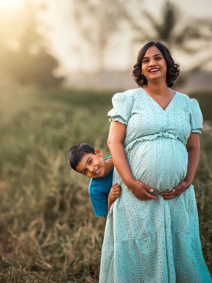 A joyful outdoor family portrait where a young boy playfully peeks out from behind his pregnant mother. The golden hour sunlight adds a warm, happy glow to this candid moment.