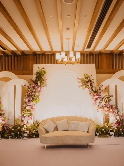 A clean and elegant stage design for an engagement ceremony. The backdrop features modern white arches adorned with delicate pink and white floral arrangements, creating a soft and romantic focal point.